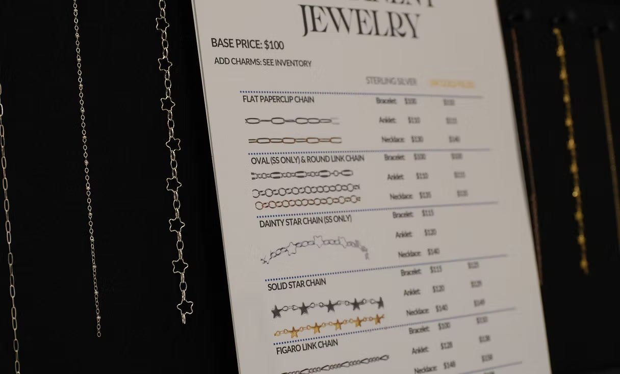 The NCG permanent jewelry price board beside sterling silver and gold chains on black display panels
