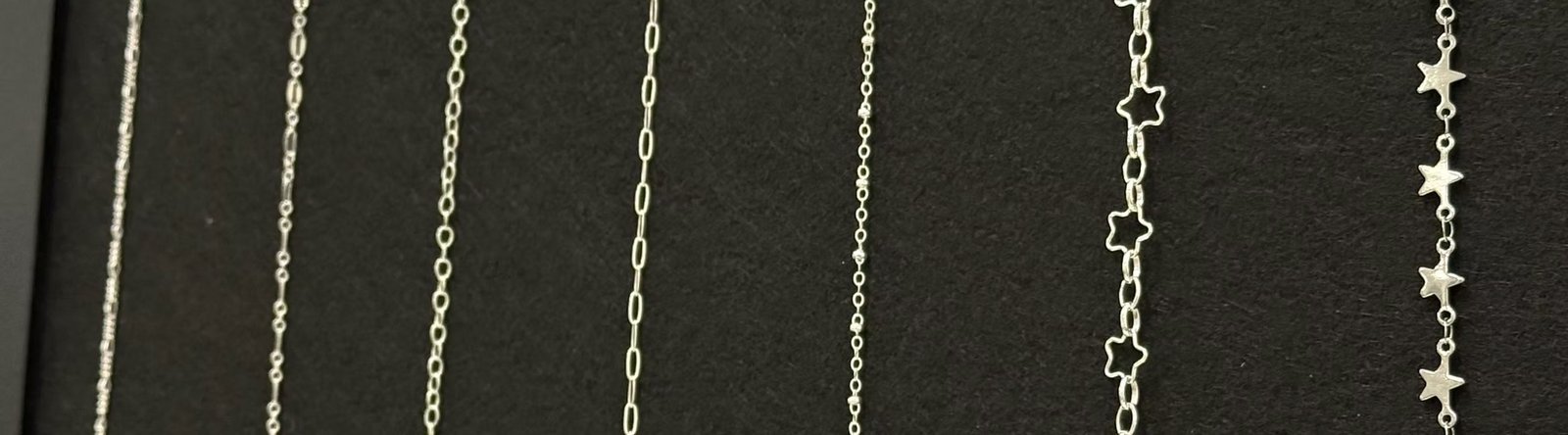 A row of sterling silver permanent jewelry chains on black felt, from fine links to open and solid stars